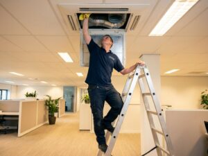 Onderhoudsmedewerker op ladder reinigt airconditioning ventilatierooster in modern kantoor met gele doek